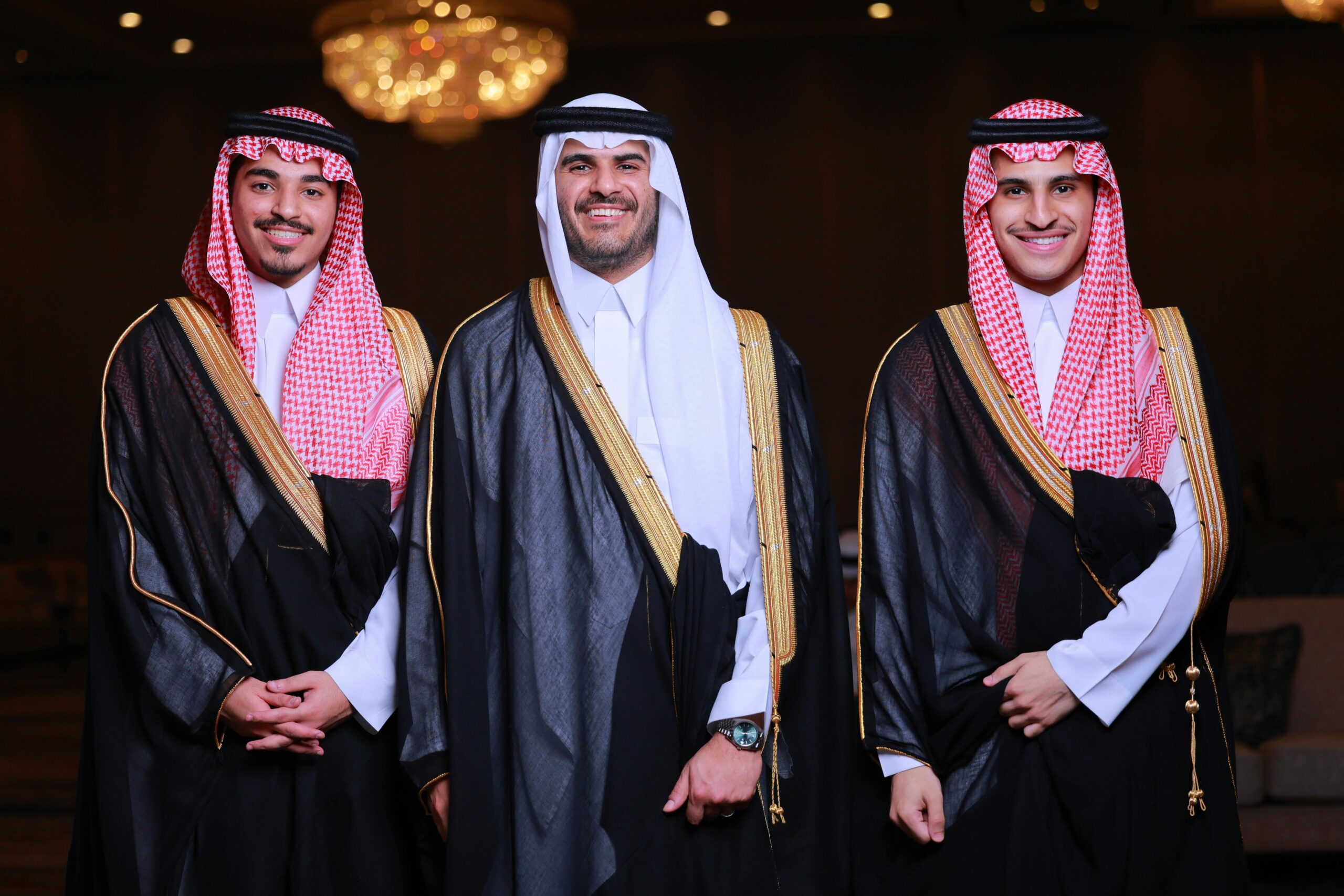 Three men in traditional Arabic robes and keffiyeh smiling at a formal indoor event.