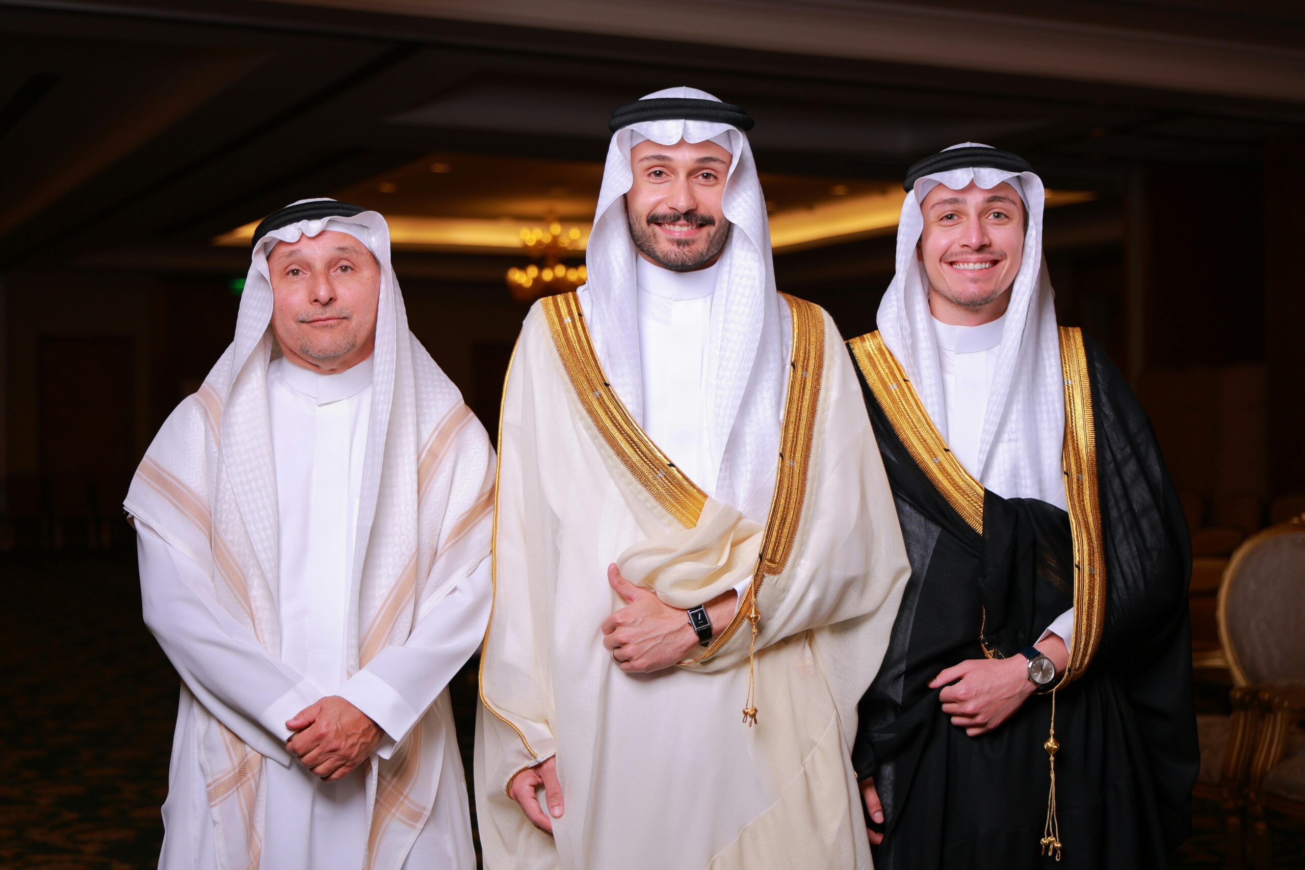 Three Middle Eastern men wearing traditional thobes indoors, smiling and posing.